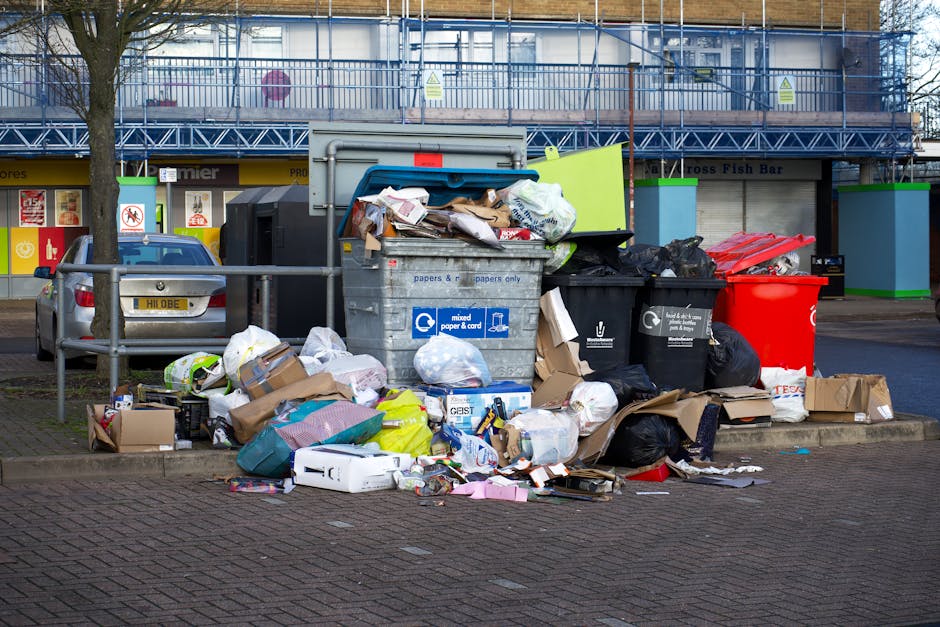 A collection of overflowing rubbish bins and scattered waste materials, including cardboard boxes, plastic bags, and paper debris, situated on a paved sidewalk in front of a commercial building. The b