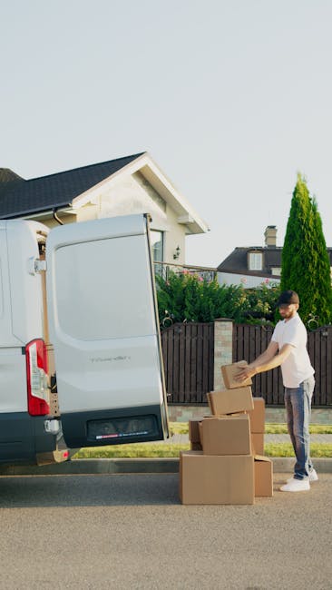 A man with a beard and wearing a cap and white shirt is seen outside through the open rear door of a moving van during a home relocation process. He is in the act of handling a cardboard moving box, w
